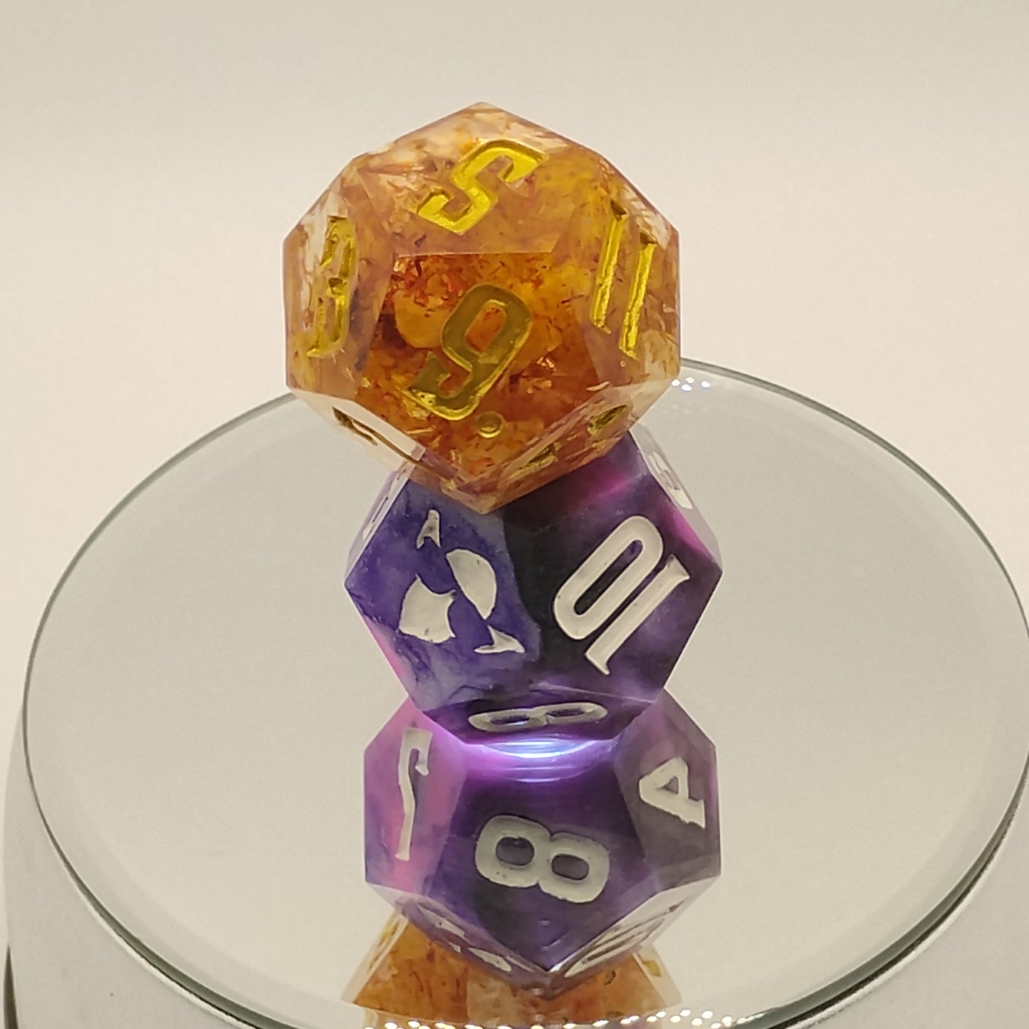 Two polyhedral dice, one orange and one purple with white numbers, on a reflective surface.