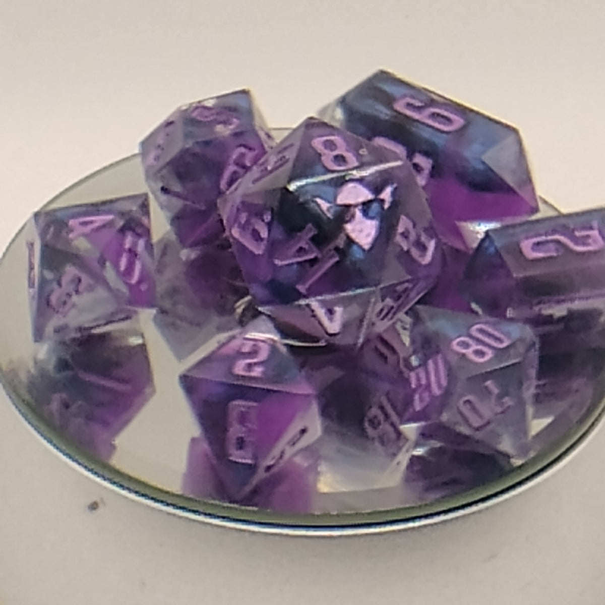 A pile of translucent purple polyhedral gaming dice with white numbering, displayed on a small reflective plate against a plain light background.